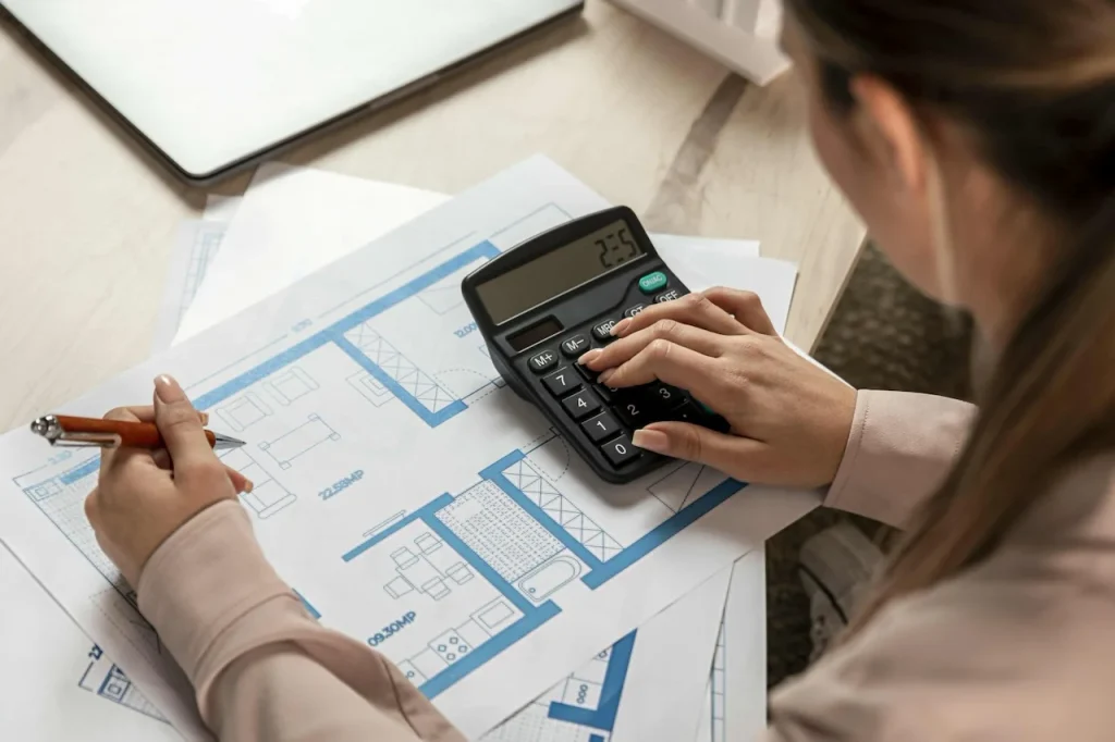Person reviewing architectural plans with a calculator while comparing estimated residential construction costs.