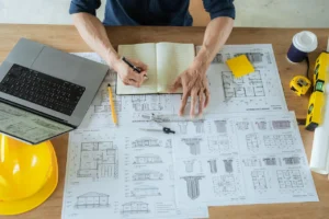 Architectural building plans, laptop, and planning documents used to organize a new home construction timeline.